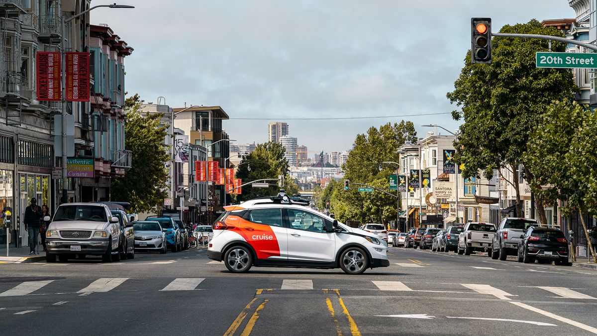A white General Motors Bolt electric vehicle with a red "Cruise" logo is crossing an intersection in downtown San Francisco, with a street of parked cars and businesses behind it.